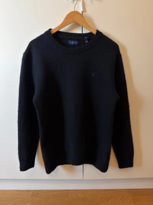 Svart stickad tröja från GANT - Klassisk svart stickad tröja från GANT med rund halsringning och diskret logga på bröstet. Tröjan har ribbstickade muddar vid ärmslut och nederkant. Perfekt för lager-på-lager och enkel att matcha med olika outfits. Passar för storlek S-M beror på kroppsvikt och form
