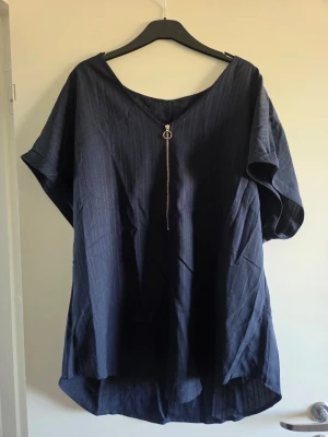 Mörkblå blus med dragkedja, XXL - En loose fit mörkblå blus med subtila ränder och kort ärm. Blusen har en snygg V-ringning framtill med dekorativ dragkedja och rundad nederkant. Perfekt för en avslappnad och trendig look.
