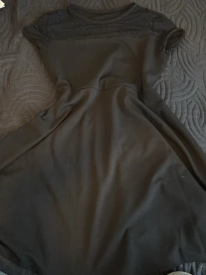 Svart kortärmad klänning med mesh - Svart klänning med korta ärmar och rund hals. Överdelen har ett transparent meshparti med diskreta prickar som ger en snygg detalj. Kjolen är utsvängd och klänningen har en feminin siluett. Perfekt för dig som gillar stilrena plagg med en twist. Kommer ej ihåg vart jag köpte från? 