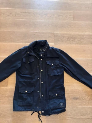 Svart vindjacka från Black Premium - Stilren svart vindjacka från Black Premium med fyra framfickor med lock och knappar, dragkedja och tryckknappar framtill samt dragsko i nederkant. Jackan har hög krage och är perfekt för blåsiga dagar.