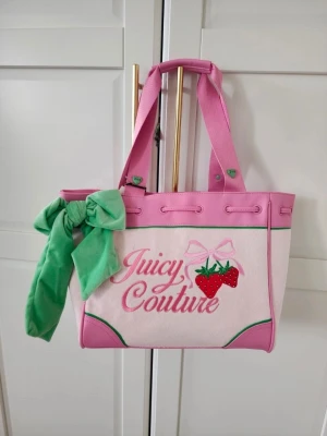 Juicy Couture shoppingväska med jordgubbar - Superfin shoppingväska från Juicy Couture i rosa och vitt med gröna detaljer. Väskan har broderade jordgubbar, en stor grön rosett och texten 'Juicy Couture' framtill. Tillverkad i tyg med rosa handtag och dekorativa sömmar.