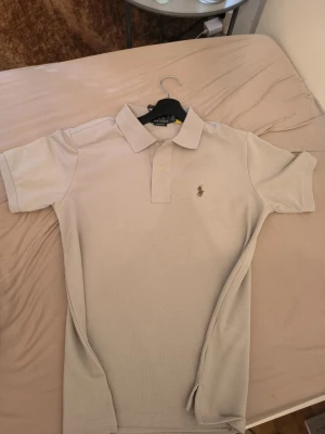 Beige pikétröja från Ralph Lauren pris kan diskuteras  - Snygg beige pikétröja från Ralph Lauren med klassisk krage och två knappar framtill. Tröjan har den ikoniska broderade loggan på bröstet. Passformen är slim och materialet känns mjukt och bekvämt, perfekt för en clean och stilren look.