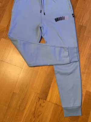 Hoodrich Mjukisbyxor Ljusblå (ny skick) - Snygga ljusblå mjukisbyxor från Hoodrich med broderad logga på ena benet. Byxorna har dragsko i midjan, muddar vid bensluten och sidofickor. Perfekta för en chill och avslappnad stil. Storlek S passar M. Nytt skick.