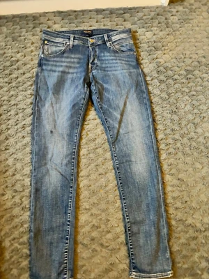 Blå slim fit jeans från Jack & Jones - Snygga blå slim fit jeans från Jack & Jones med klassisk femficksdesign och guldfärgade detaljer vid knappen. Jeansen har en lätt tvättad look och raka ben, perfekta för en avslappnad stil.