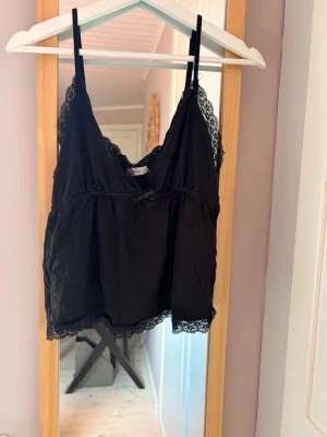 Svart spetslinne med tunna axelband - Säljer ett svart linne med tunna axelband och spetsdetaljer längs urringning och nederkant. Linnet har en liten rosett framtill och en lös, bekväm passform. Perfekt att styla under en skjorta eller som det är.