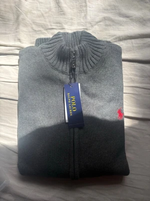 Grå zip-tröja från Polo Ralph Lauren - Snygg grå zip-tröja från Polo Ralph Lauren med ribbad krage och dragkedja framtill. Tröjan har ett diskret rött broderat logomärke på bröstet och är stickad i mjukt material. Perfekt för en chill och stilren look. Helt ny med tag kvar! Hör av er vid eventuella frågor!
