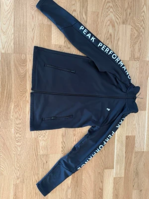 Svart tröja från Peak Performance - Svart tröja från Peak Performance med vit logga på bröstet och text längs ärmarna. Tröjan har två dragkedjeförsedda fickor framtill och hel dragkedja. Sportig och stilren design med lång ärm och hög krage.