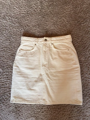 Vit jeanskjol med fickor - Snygg vit kort jeanskjol med klassiska fem fickor och knapp framtill. Kjolen har en rak passform och är tillverkad i bomull, perfekt för en fräsch och stilren look. Passar till många olika outfits och är enkel att matcha.