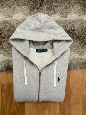 Ljusgrå Polo Ralph Lauren Zip Up Hoodie - Storlek S - Hej! 👋 Säljer en riktigt stilren ljusgrå Polo Ralph Lauren zip-up hoodie i storlek S. Den har den klassiska mörkblå pony-loggan broderad på bröstet – en klockren färgkombination! 🐎 Tröjan är i mycket fint skick och känns väldigt fräsch. 100% äkta med tvättlappar kvar (ingen QR-kod på denna modell). En mångsidig tröja som passar perfekt som basplagg året runt. Fraktar snabbt! 🚛📦💬💯 
