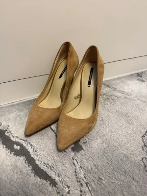 Beige mockapumps från Zara - Snygga pumps i beige mocka från Zara med spetsig tå och hög klack. Skorna har en elegant siluett och gulddetalj bak på hälen som ger en exklusiv touch. Perfekta för dig som vill ha en stilren och trendig look.