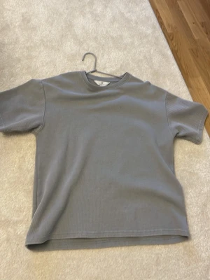 Grå ribbad t-shirt från H&M - Säljer en stilren grå t-shirt från H&M med ribbad struktur. T-shirten har korta ärmar och rund halsringning, perfekt för en avslappnad och trendig look. Materialet är mjukt och bekvämt, passar till många olika outfits.