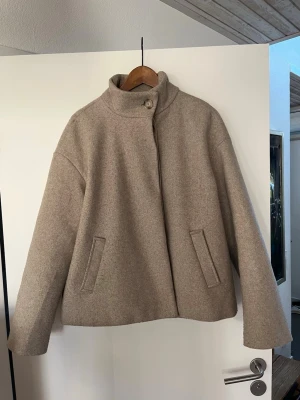 Beige kappa med krage - Stilren beige kappa med rak passform och hög krage. Kappan har knäppning framtill med fyra knappar och två snedställda fickor. Nypris 1000kr