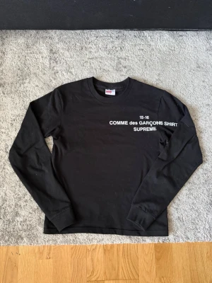 Svart långärmad topp Supreme x Comme des Garçons - Svart långärmad topp från Supreme och Comme des Garçons med vit text på bröstet. Klassisk rund halsringning och relaxed fit. Perfekt för dig som gillar streetwear och vill sticka ut med en ikonisk collab.
