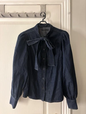 Mörkblå jeansblus med knyt från Stockh lm - Snygg mörkblå jeansblus från Stockh lm med knytband i halsen och puffiga axlar. Blusen har långa ärmar, knappar framtill och är gjord i ett mjukt denimtyg. Perfekt för dig som gillar en trendig och avslappnad look. Endast använd 1 gång