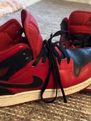 Nike Air Jordan 1 Retro High röd/svart - Klassiska Nike Air Jordan 1 Retro High sneakers i rött och svart läder med svart swoosh och vingar-logga på sidan. Skorna har högt skaft, svart snörning och vit mellansula. Perfekta för dig som gillar ikoniska och sportiga sneakers med streetkänsla.
