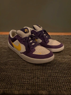 Nike SB sneakers lila och gula detaljer - Nike SB sneakers i lila och vitt med gula swoosh-detaljer. Ovandelen är i mocka och textil för en skön och snygg look. Använda typ tre gånger bara. Storleken är 46 men jag skulle säga att dom är små i storleken då de passar mig med 45 vanligtvis
