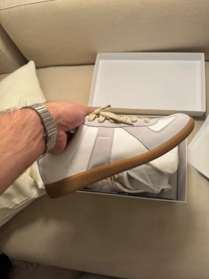 Margiela Gats - Stilrena vita sneakers med beige mockadetaljer och beige snören. Skorna har en klassisk låg siluett och en brun gummisula som ger en retrovibe. Perfekta för dig som gillar minimalistisk och tidlös design. Materialet är en mix av läder och mocka.