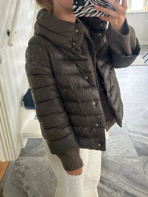 Mörkbrun hernojacka med krage - Snygg mörkbrun dunjacka med hög krage. Västen har tryckknappar framtill och en lätt glansig yta. Perfekt att slänga över en stickad tröja för extra värme. Enkel och stilren modell som passar till många outfits. Storlek 40, skulle säga att den är i en S-M 