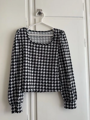 Svartvit rutig långärmad topp - Trendig långärmad topp med svartvitt rutigt mönster och rundad urringning. Modellen är croppad och har breda muddar vid ärmsluten. Perfekt för dig som vill sticka ut med en klassisk men edgy stil.