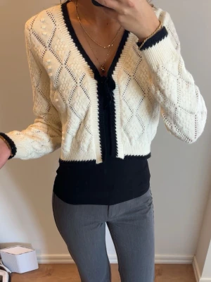 Vit stickad kofta med svarta detaljer - Snygg vit stickad kofta med svarta kanter och rosett framtill. Koftan har ett  mönster i diamantform. Perfekt att slänga över ett linne eller t-shirt för en trendig look.