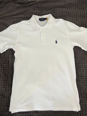 Vit pikétröja från Polo Ralph Lauren - Klassisk vit pikétröja från Polo Ralph Lauren med broderad marinblå logga på bröstet. Tröjan har korta ärmar, krage och knäppning med två knappar framtill. Perfekt för en stilren och tidlös look.