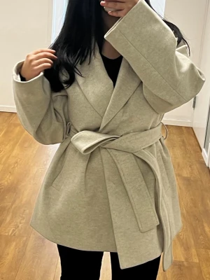 Beige kappa med knytskärp - Trendig beige kappa med bred krage och knytskärp i midjan. Kappan har en rak och avslappnad passform med stora slag och långa ärmar. Perfekt för lager-på-lager och ger en stilren look. Materialet är mjukt och känns lyxigt.