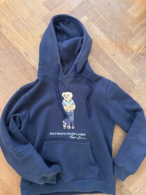 Marinblå hoodie Polo Ralph Lauren - En Polo RL bear hoodie med vintage stil. Notera att det som ser ut som rivmärken är en del av hoodien för den ska se lite vintage ut.