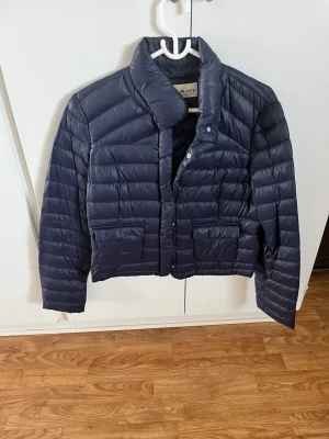 Mörkblå pufferjacka från de.corp - Snygg mörkblå pufferjacka från de.corp Esprit Urban Casual. Jackan har en kortare modell, hög krage och två framfickor. Quiltad design med horisontella sömmar och tryckknappar framtill. Perfekt för kalla vinterdagar.