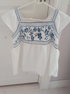 Vit blus med blå broderi och knyt - Supersöt vit blus med blått broderat mönster framtill och dekorativa axelband. Toppen har volangärmar och knyt i ryggen för extra detalj. Perfekt för en somrig och fräsch look. Den är lite större i varianten skulle mer säga att den är som en M