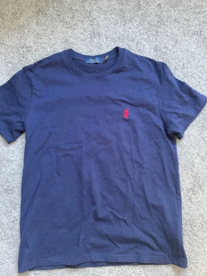 T-shirt Ralph Lauren - Klassisk marinblå t-shirt från Polo Ralph Lauren med röd broderad logga på bröstet. T-shirten har rund halsringning och korta ärmar. Tillverkad i mjuk bomull för en skön känsla och enkel stil.