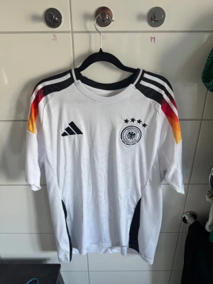 Tysklands matchtröja Adidas herr - Vit matchtröja från Adidas med Tysklands landslagsmärke och fyra stjärnor på bröstet. Tröjan har svarta, röda och gula detaljer på axlarna, samt svarta ränder och meshpaneler på sidorna. Tillverkad i lätt och ventilerande material, perfekt för fotboll.