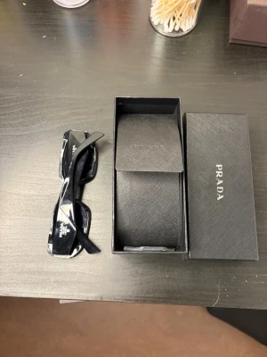 Svarta solglasögon från Prada - Snygga svarta solglasögon från Prada med rektangulära bågar och tydlig logga på sidorna. Kommer med originaletui och ask. Perfekta för dig som vill ha en stilren och lyxig accessoar till din look.