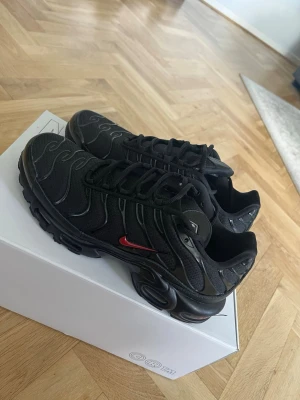 Nike Air Max Plus svarta sneakers - Svarta Nike Air Max Plus sneakers med mesh- och syntetmaterial, ikoniska vågformade detaljer och en liten röd Swoosh på sidan. Skorna är nytt skick, har provat dom 2-3 gånger men passade inte. Nypris 2649kr