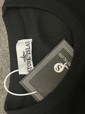 Svart sweatshirt från Stone Island - Säljer en svart sweatshirt från Stone Island med klassisk rund hals och ribbade muddar vid ärmslut och nederkant. Tröjan har den ikoniska Stone Island-loggan på vänster ärm. Tillverkad i mjukt bomullsmaterial för en clean och stilren look.