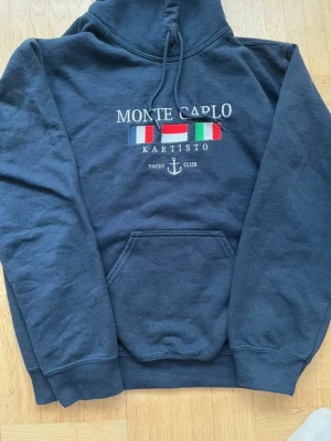 Monte Carlo Kartisto hoodie - Svart hoodie från Kartisto med broderad text 'Monte Carlo' och färgglada flaggor på bröstet. Har stor huva, dragsko och magficka. Materialet är mjukt och skönt, perfekt för en avslappnad stil. Broderad ankare och 'Yacht Club' ger en nautisk vibe.