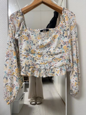 Blommig blus från Bik Bok - Supersöt blommig blus från Bik Bok med volangkant nertill och smockad byst. Blusen har långärmade puffärmar med resår vid ärmslut och en fyrkantig ringning. Tunt, luftigt tyg i vitt med gula, blå och lila blommor.