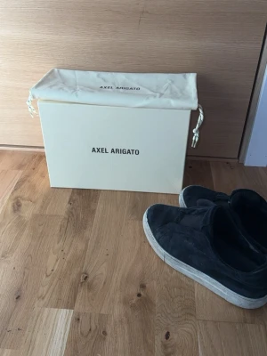 Svarta slip-on sneakers Axel Arigato - Svarta slip-on sneakers från Axel Arigato i mocka med vit, tjock sula. Snygg minimalistisk design utan snörning, perfekt för en clean och stilren look. Skorna levereras med originalkartong och dustbag.