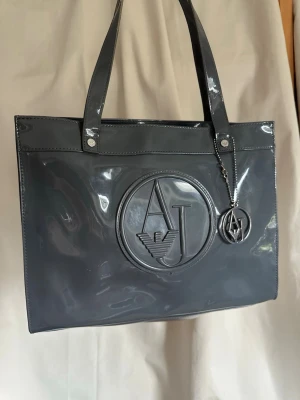  Armani Jeans Väska - Snygg grå glossy väska från Armani Jeans i glansigt lackmaterial. Väskan har dubbla axelband, stort AJ-logotyp framtill och en matchande logotypberlock i metall. Perfekt för dig som vill ha en stilren och trendig accessoar med plats för allt du behöver.