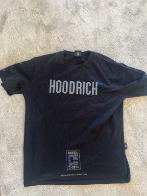 Svart HOODRICH t-shirt med tryck - Svart t-shirt från HOODRICH med stort vitt och blått tryck på bröstet och en stor grafisk print på ryggen. Rund halsringning och korta ärmar. Materialet är mjukt bomullstyg, perfekt för en streetwear-look.