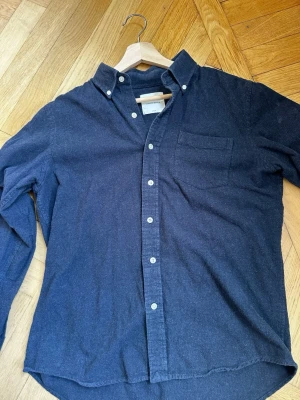 Mörkblå skjorta från A Day's March - Klassisk mörkblå skjorta från A Day's March i mjuk bomull. Skjortan har button-down krage, bröstficka och vita knappar framtill. Perfekt för en stilren och avslappnad look. Lite liten i storlek. Närmare storlek S.