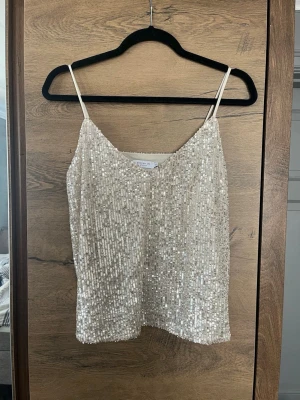 Glittrig paljettopp med smala band - Snygg glittrig topp med smala axelband och v-ringning. Toppen är täckt av champagne paljetter och har en croppad passform. Perfekt för fest eller nyår. Jag har sytt in den lite i båda sidor högst upp och sytt ner banden någon centimeter för en bättre passform. Om man inte vill ha det så är det väldigt enkelt att sprätta upp så får man den som vanligt utan skador igen. Använd en gång så väldigt bra skick.