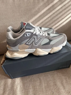 New Balance 9060 grå sneakers - Stilrena New Balance 9060 sneakers i olika nyanser av grått och beige med vita detaljer. Ovandelen är i mesh och mocka, med en chunky sula i beige och svart. Ikoniska N-loggan på sidan och röd detalj på plösen. Perfekta för dig som gillar streetwear och retrostil.