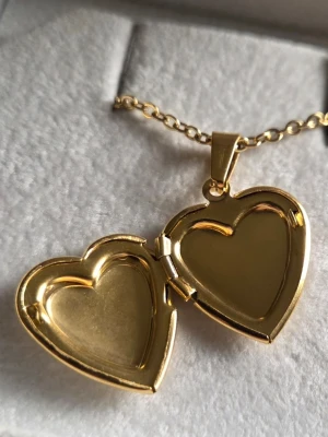 Guldigt hjärthalsband med medaljong - Snyggt halsband i guldfärgat rostfritt stål med ett hjärtformat medaljonghänge. Medaljongen går att öppna och har plats för små bilder eller minnen. Utsidan är dekorerad med fina snirkliga detaljer runt hjärtat. Perfekt accessoar för en personlig touch.