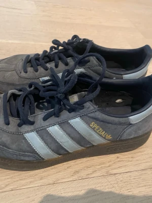 Adidas Spezial blå sneakers - Klassiska Adidas Spezial sneakers i blå mocka med ljusblåa ränder och guldfärgad text på sidan. Skorna har mörkblå snören och en brun sula. Perfekta för dig som gillar retrostil och streetwear. Storlek 40 2/3 