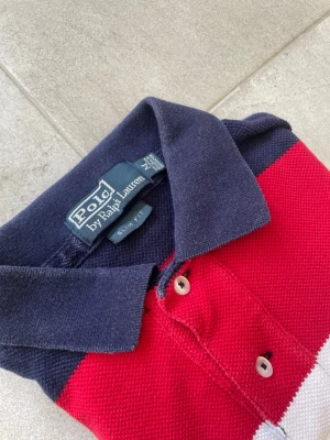Randig pikétröja från Polo Ralph Lauren - Snygg pikétröja med breda ränder i rött, vitt, ljusblått och marinblått från Polo Ralph Lauren. Tröjan har korta ärmar, krage och knappar framtill. Slim fit-modell med klassisk logga broderad på bröstet.                                               Mått 📏 Längd från axel: 67cm Axelbredd: 42cm Skador 🔧: Inga  Storlek 🧍: M  Jag kan förhandla mig till lägre pris.🙋💵 Om du har några frågor, vänligen skriv till mig 🤗