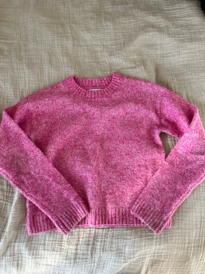 Rosa stickad tröja med rund hals - Mysig rosa stickad tröja med rund hals och långa ärmar. Tröjan har ribbade muddar vid ärmslut och nederkant. Perfekt för att hålla värmen under kyliga dagar och ger en färgklick till din outfit.