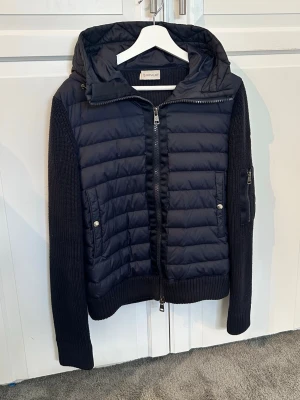 Svart pufferjacka från Moncler - Snygg svart pufferjacka från Moncler med stickade ärmar och vadderad front. Jackan har huva, dragkedja framtill och fickor med tryckknappar. Ärmarna har en stickad struktur och en ficka med dragkedja på ena ärmen. Perfekt för kalla dagar. Som ny. Tar bara plats. Ställ gärna frågor 😁😌