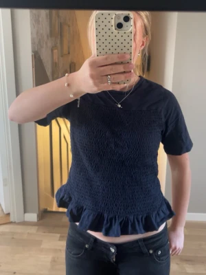 Mörkblå smockad topp med volangkant - Supersöt mörkblå topp med smockad framsida och volangkant nedtill. Toppen har korta ärmar och rund halsringning. Perfekt att styla med jeans eller kjol för en trendig look.