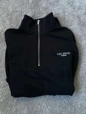 Svart half-zip hoodie från Les Deux - Snygg svart hoodie från Les Deux med halv dragkedja framtill och broderad logga på bröstet. Tillverkad i mjukt material som känns skönt mot huden. Perfekt för dig som gillar stilrena och enkla plagg med en modern touch. Använd oerhört få gånger (2-3) då jag växte ur den några veckor efter jag fått den.