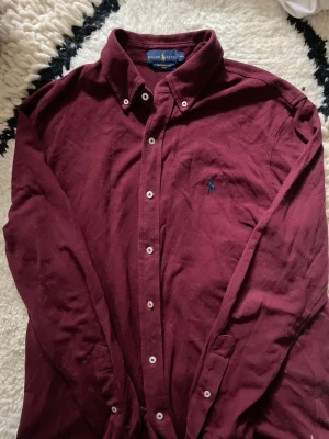 Vinröd skjorta från Ralph Lauren - Vinröd långärmad skjorta från Ralph Lauren med klassisk krage och knappar framtill. Skjortan har broderad logga på bröstet och är tillverkad i mjuk bomull. Perfekt för en stilren och avslappnad look.